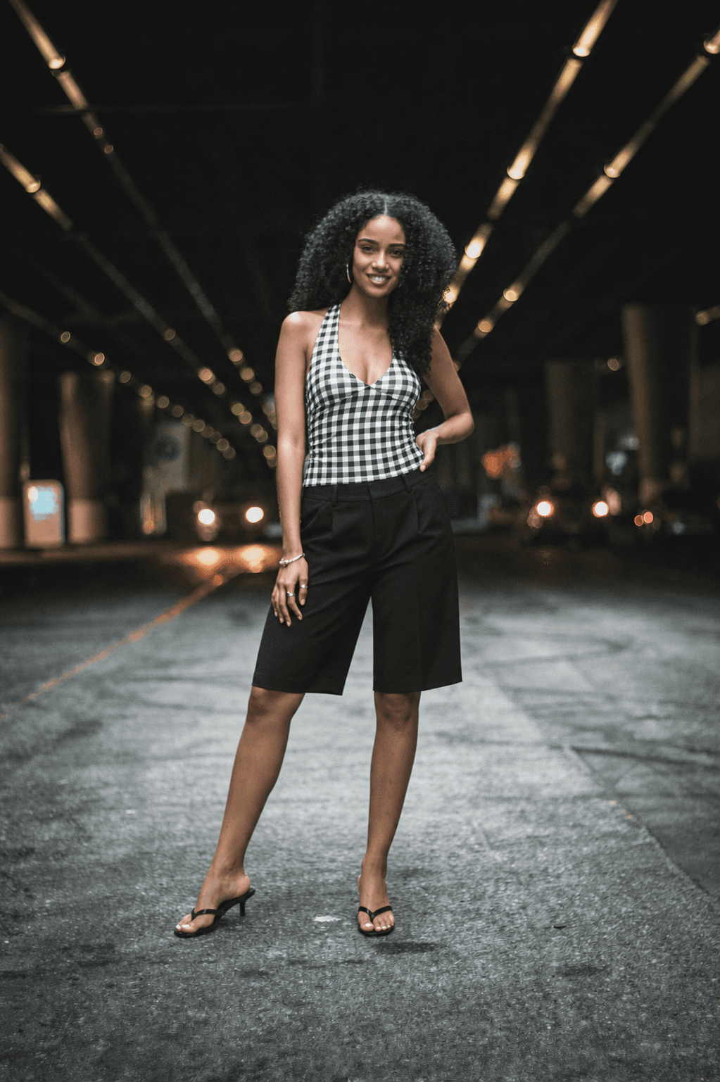 The same supplied real woman model in the same photo, wearing the reference outfit: a gingham halterneck top, black tailored Bermuda shorts, and black kitten-heel mules